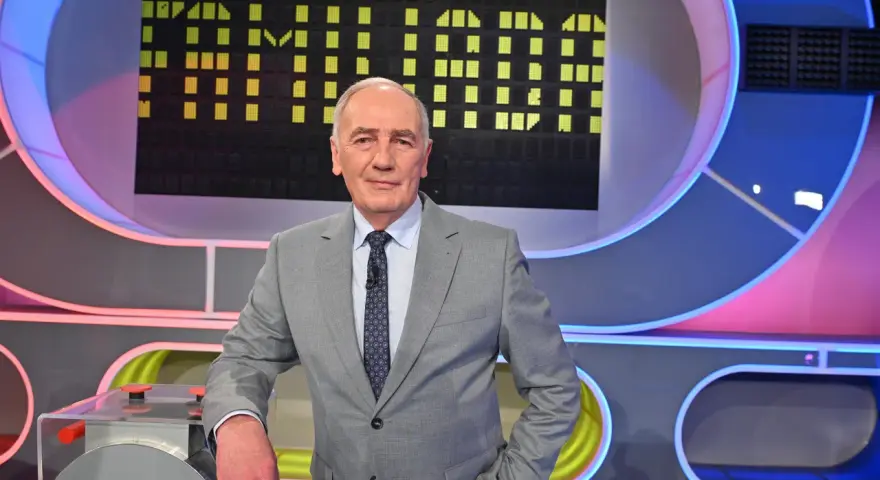 Mężczyzna w garniturze na tle planszy teleturniejowej, wyglądający na siłach i gotowy do gry, by być zdrowym jak Familiada.