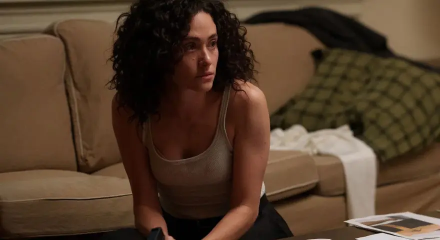 Kobieta z kręconymi włosami, w podkoszulku, siedzi na podłodze, trzymając pistolet. Scena z filmów i seriali Emmy Rossum.