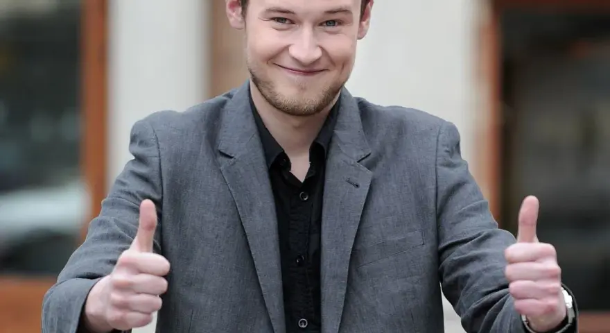 Krzysztof Wójcik z "Milionerów" pokazuje kciuki w górę, uśmiechając się.