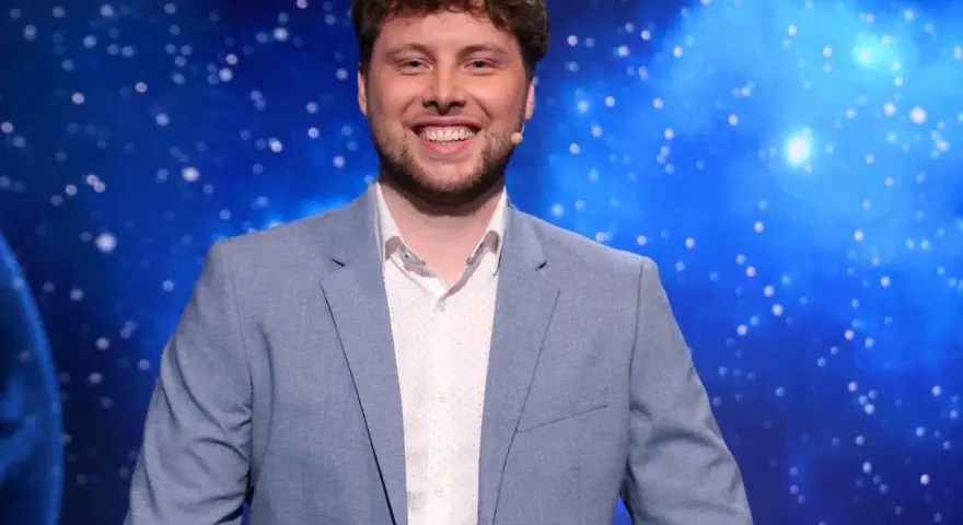Uśmiechnięty mężczyzna w jasnej marynarce, jeden z dziesięciu jak się zgłosić, na tle kosmicznego nieba.