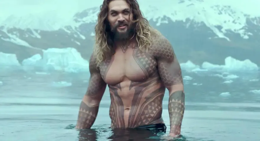 Jason Momoa w wodzie, z tatuażami, jak w filmach i serialach. W tle góry i lód.