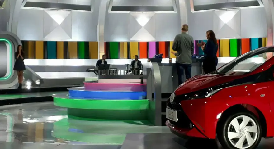 Studio telewizyjne, gdzie nagrywane jest koło fortuny. Widać scenografię z kolorowymi panelami, schodami i czerwonym samochodem.