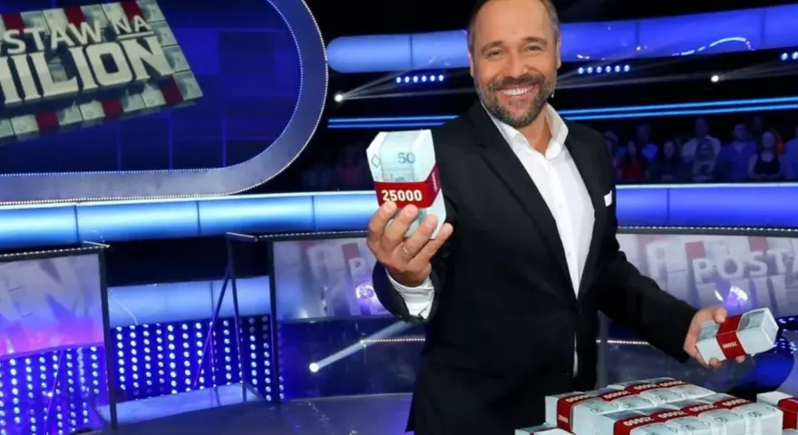 Uśmiechnięty prowadzący teleturniej trzyma w dłoni paczkę pieniędzy. Na stole piętrzą się stosy banknotów.