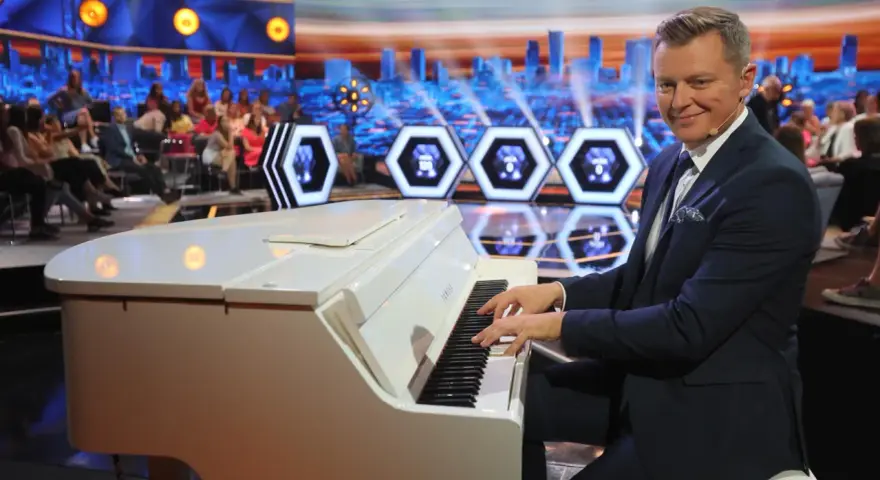 Mężczyzna gra na białym pianinie w studio telewizyjnym. Czy to melodia z programu "Jaka to melodia"?