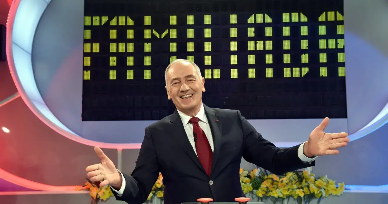 Uśmiechnięty mężczyzna w garniturze, który prowadzi Familiadę, gestykuluje radośnie.