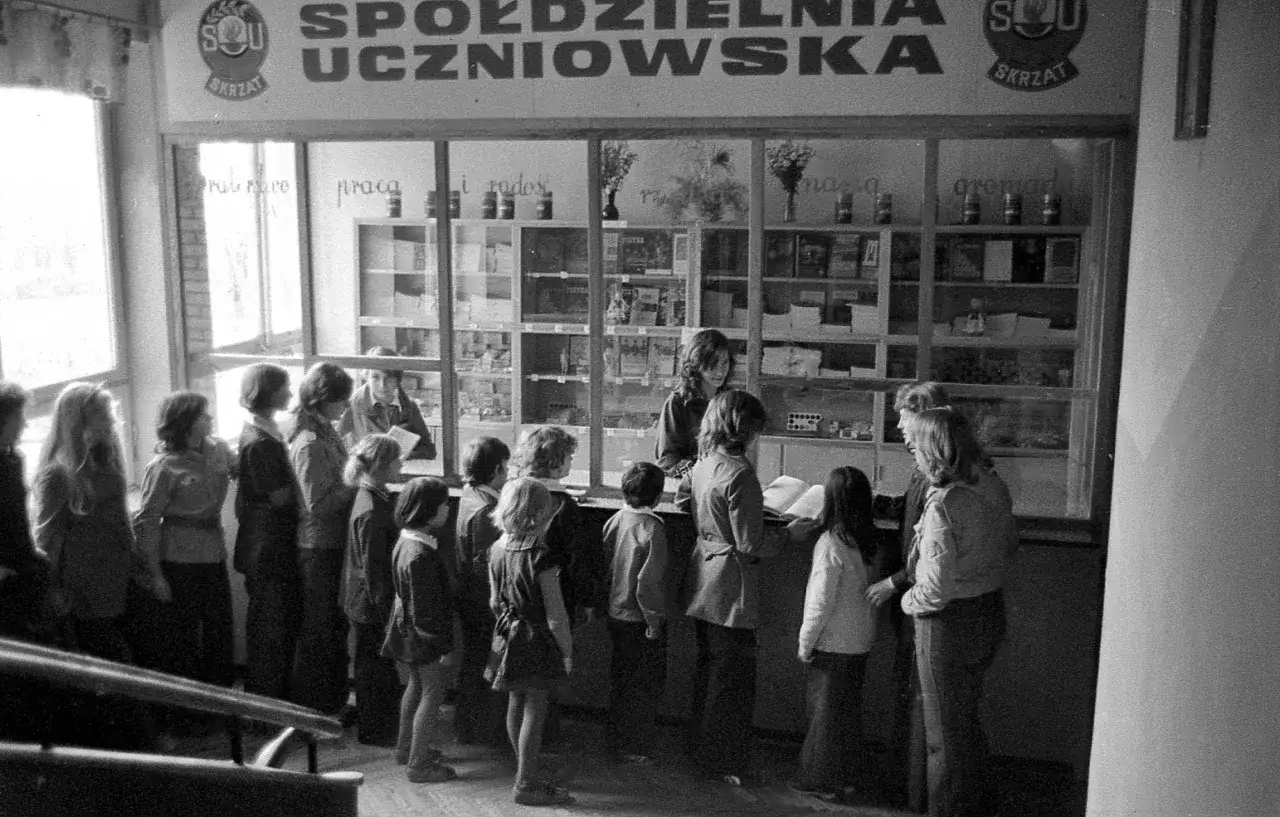 Dzieci stoją w kolejce do sklepiku szkolnego, gdzie można kupić zeszyty, książki i inne artykuły.