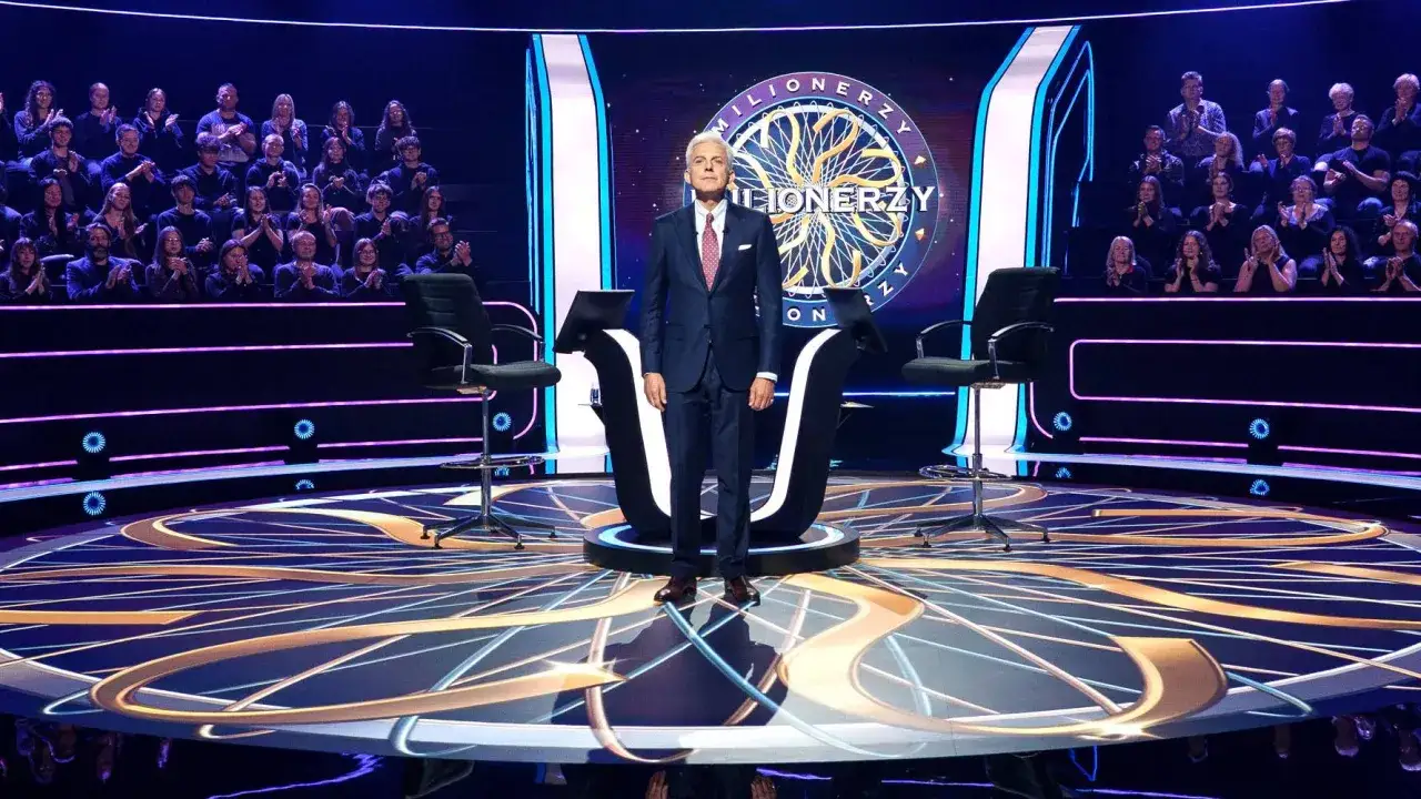 Hubert Urbański na scenie Milionerów. Widownia w tle. Koła ratunkowe w Milionerach to: 50:50, telefon do przyjaciela, pomoc publiczności.