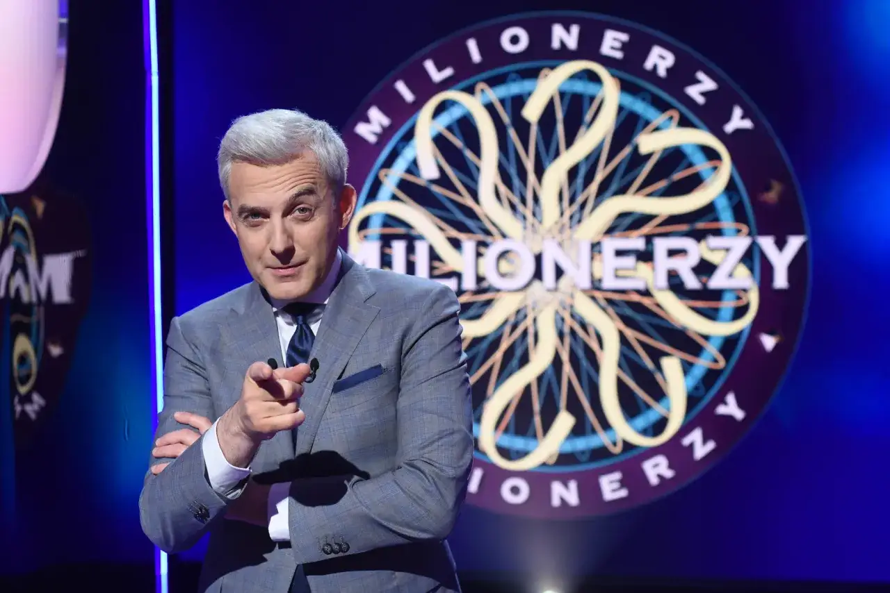 Prowadzący Hubert Urbański w garniturze wskazuje palcem, a za nim logo teleturnieju