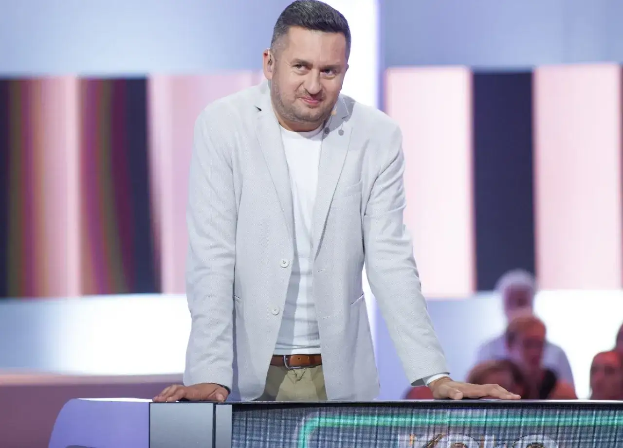 Mężczyzna w jasnej marynarce, który prowadzi Koło Fortuny, z uśmiechem spogląda na widownię.