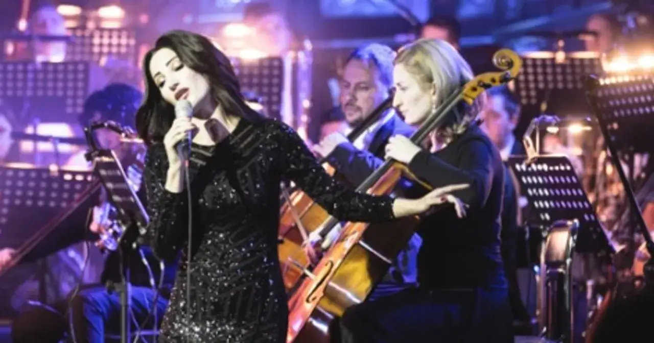 Wokalistka w błyszczącej sukni śpiewa z mikrofonem, a obok niej gra wiolonczelistka. Koncert z udziałem polskich piosenkarzy i orkiestry.