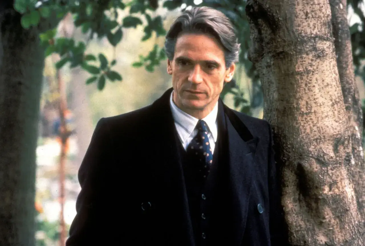 Jeremy Irons w eleganckim płaszczu i krawacie, jak z najlepszych jeremy irons filmy. Stoi oparty o drzewo, w otoczeniu zieleni.