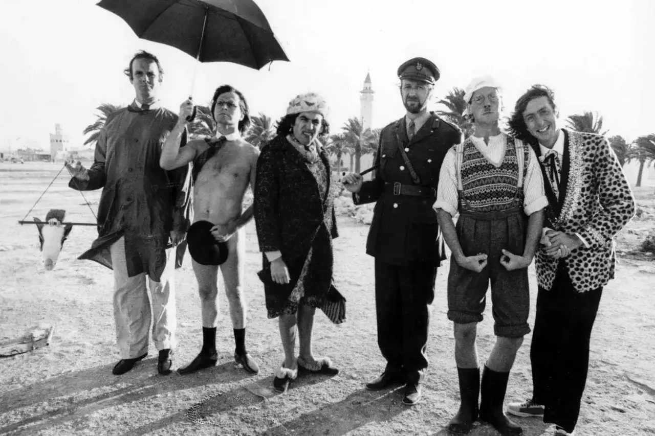 Monty Python w absurdalnych strojach, z krową na patyku i parasolem, pozuje do zdjęcia.