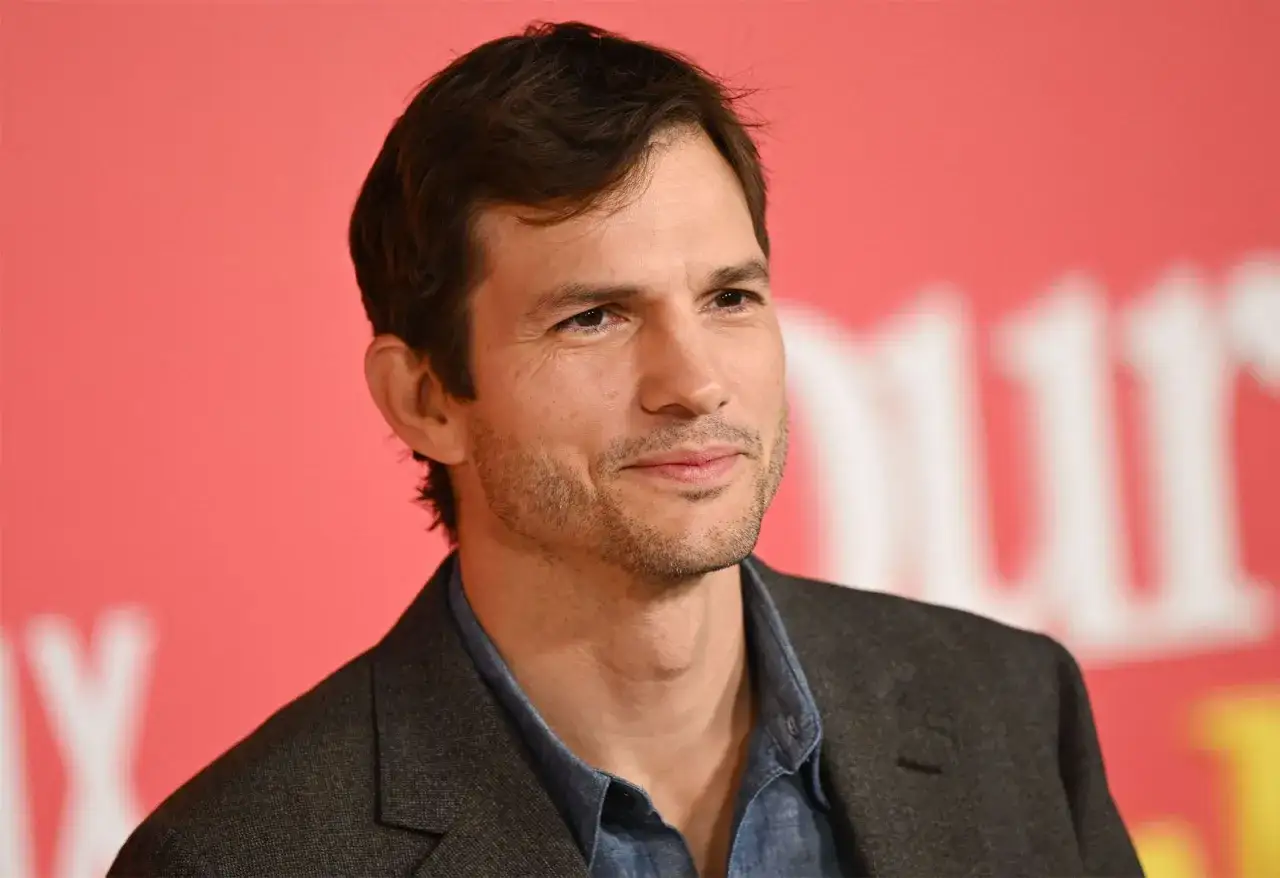 Ashton Kutcher, znany z filmów, seriali i programów, uśmiecha się lekko na tle różowej ściany.