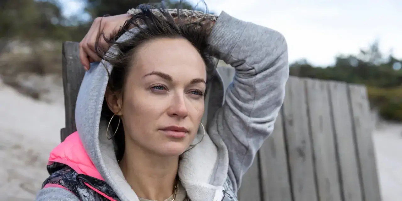 Pati 2, z ręką na głowie, w szarej bluzie z kapturem, siedzi na plaży.