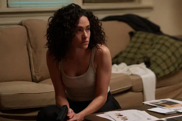 Kobieta z kręconymi włosami, w podkoszulku, siedzi na podłodze, trzymając pistolet. Scena z filmów i seriali Emmy Rossum.