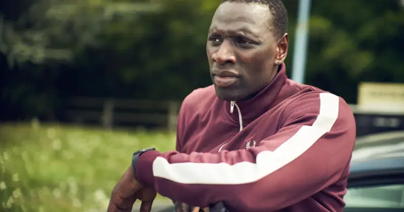 Omar Sy w bordowej bluzie z białymi pasami, gotowy do akcji w kolejnych filmach, serialach i programach.