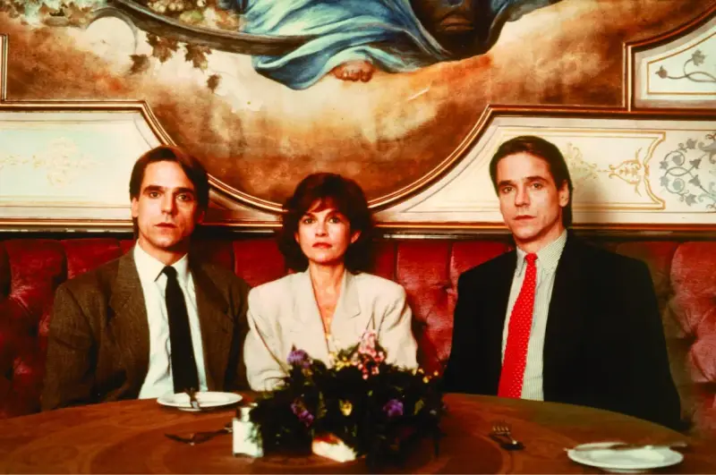 Jeremy Irons w filmach, tutaj z dwiema kobietami, siedzi przy stole w eleganckiej restauracji.